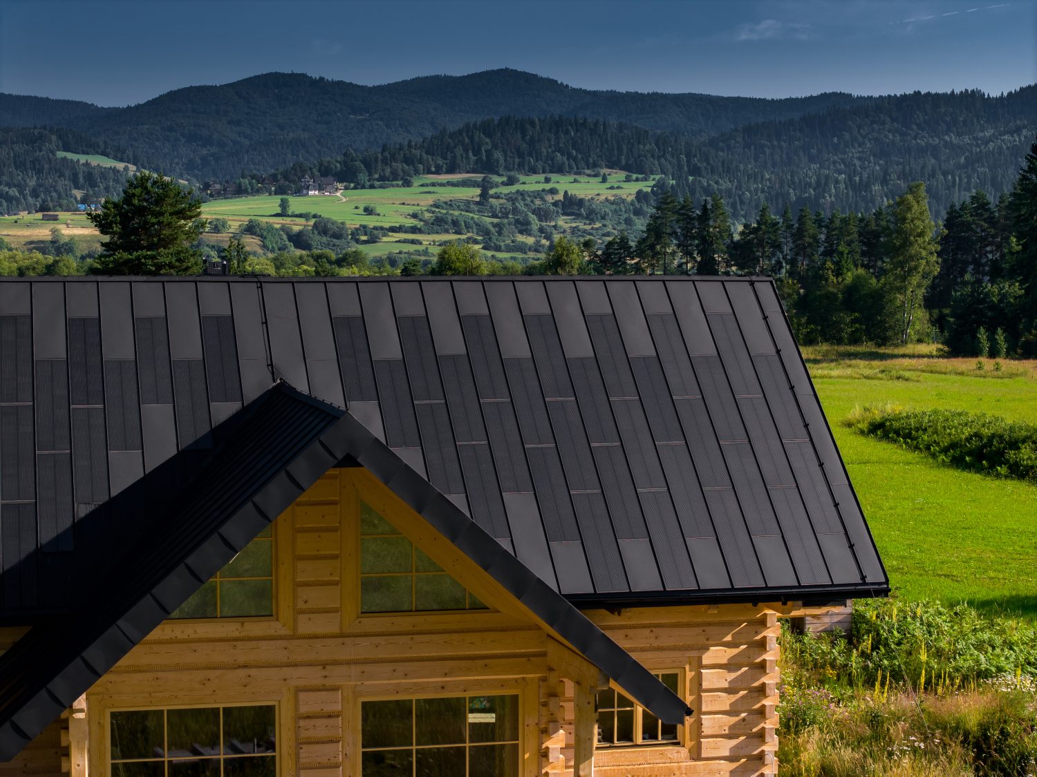 Log house with a dark SolRoof FIT VOLT standing-seam solar roof, seen from above; manufacturer photo of a European installation